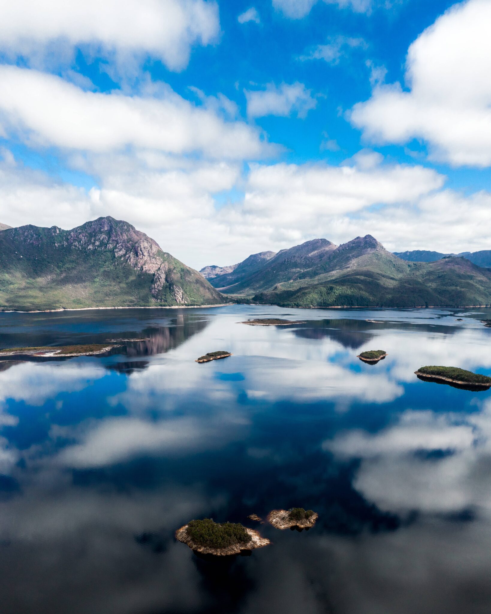 A serene landscape featuring the calm Dove Lake in Cradle Mountain reflecting the blue sky and scattered white clouds. Surrounding the lake are lush green mountains with rocky peaks.