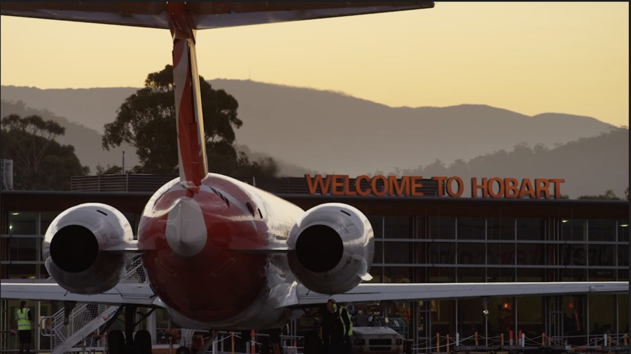 Rear of plane approaching airport terminal, Welcome to Hobart sign above building, trees and mountains in background.
