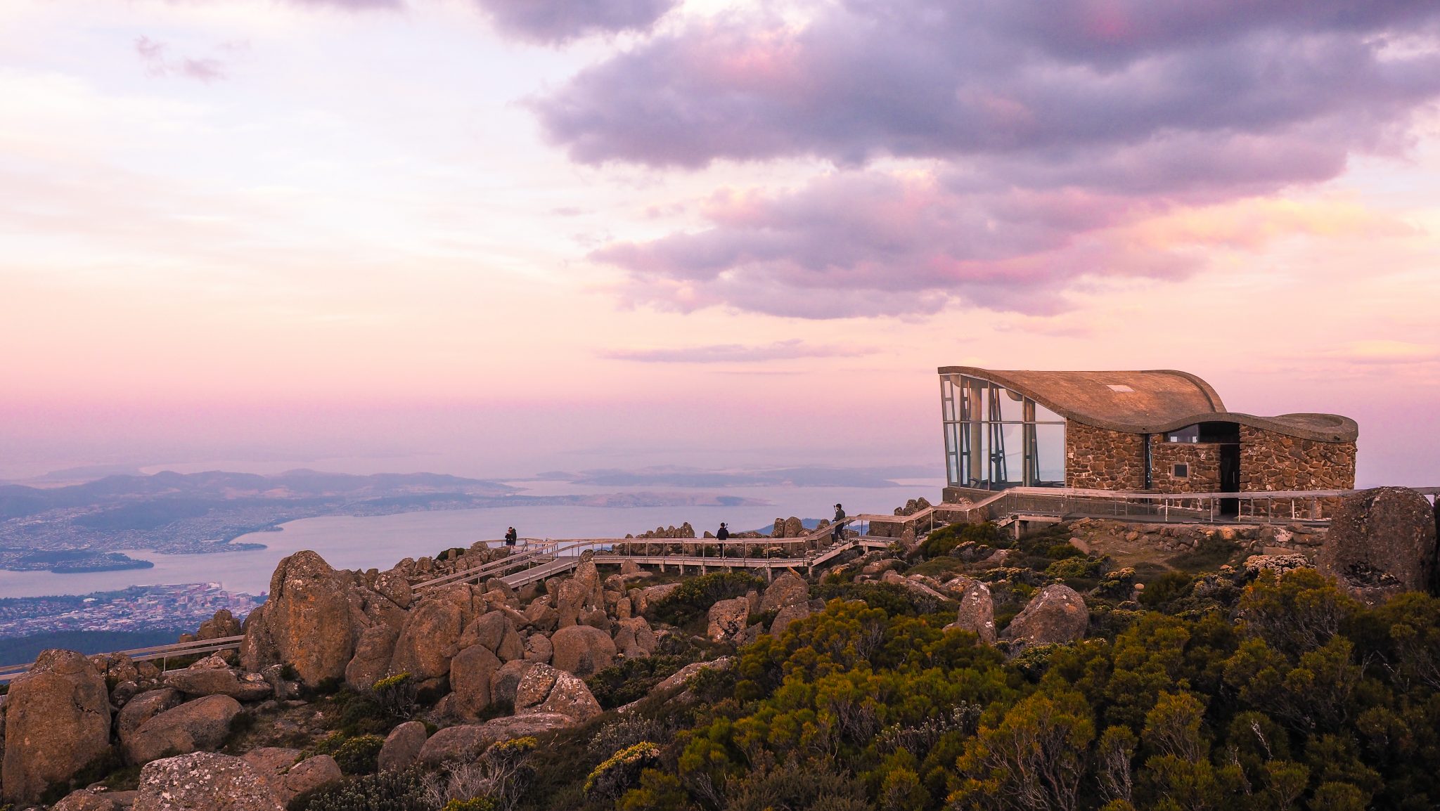 Mount Wellington kunanyi observation building from mountain summit, large rocks in foreground, overlooking Hobart and river, dusky purple and yellow sunrise.