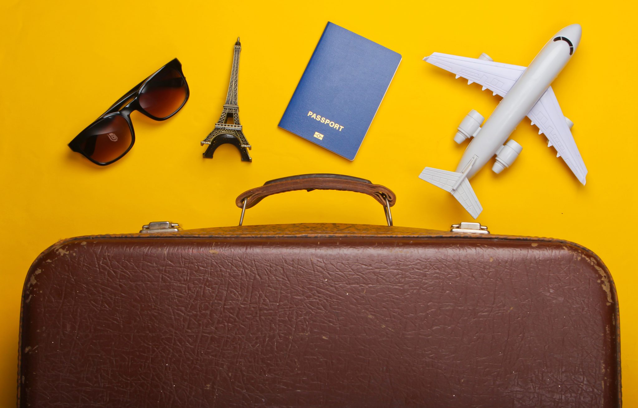 Brown suitcase, sunglasses, Eiffel Tower ornament, passport and model airplane, on yellow background.