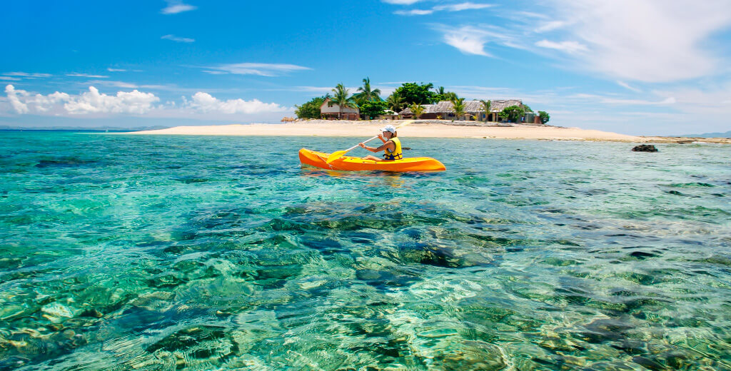 Small tropical island with person in orange canoe, and clear waters and underwater coral in foreground.