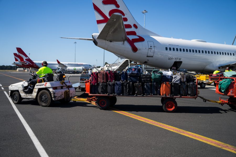 Baggage handling vehicle loaded with suitcases, driving past Virgin Australia plane.