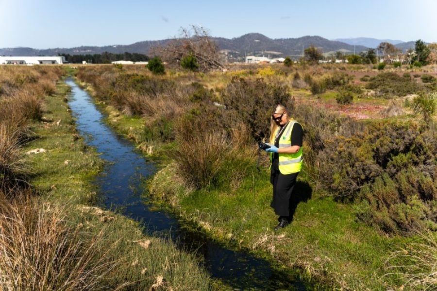 Female airport staff with green high vis jacket standing on natural landscape besides a water stream.