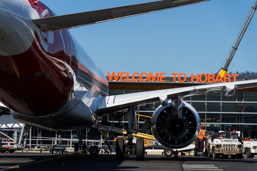 Close-up of the wing and engine of a parked airplane at the airport, with a Welcome to Hobart sign in orange letters above. Ground crew and equipment are visible near the aircraft.