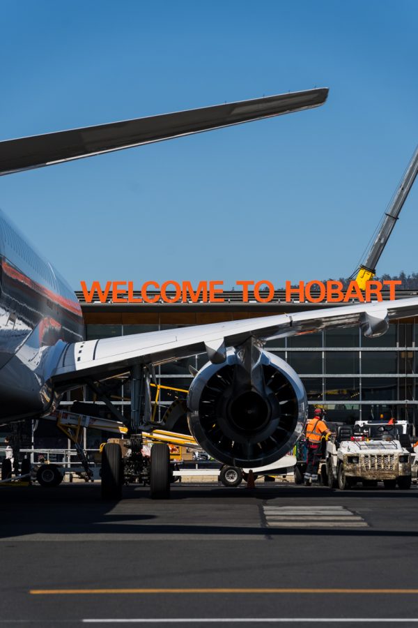 Close-up of an airplane's engine and landing gear on the tarmac. A building in the background displaying an orange sign reading 'WELCOME TO HOBART.