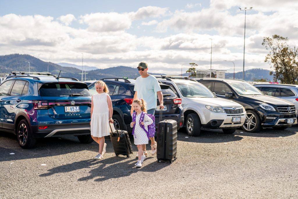 Father and two girls walking through carpark, pulling suitcases.