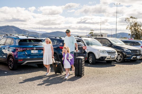 Father and two girls walking through carpark, pulling suitcases.