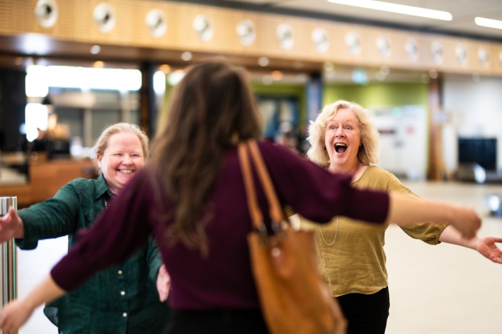 Two women with open arms and smiles approaching a third woman, also with open arms, arriving into the airport.