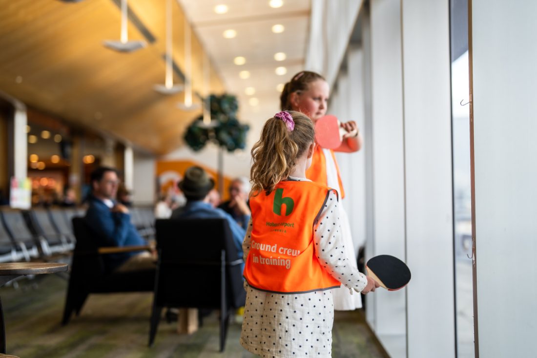 Two girls wearing orange visible jackets and holding on to table tennis rackets while watching out into the airside.