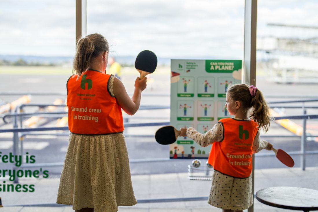 Two girls in orange high vis jackets watch over the airside of the airport holding onto table tennis rackets.