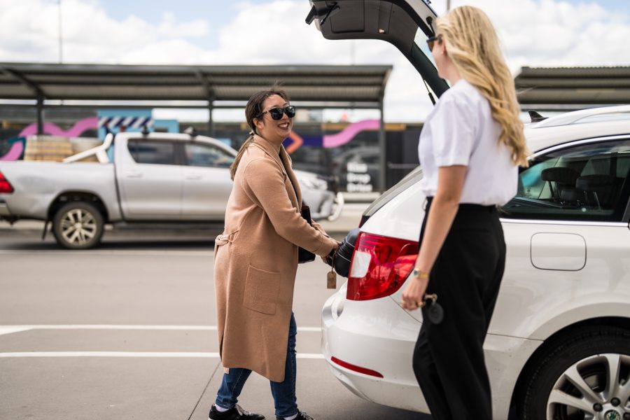 Smiling woman loading bag into parked car at airport, being greeted by another woman.