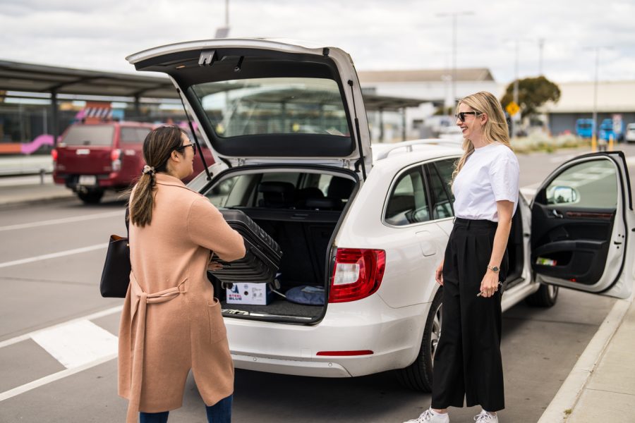 Two women talking at parked car at airport, while loading a bag into the boot of car.