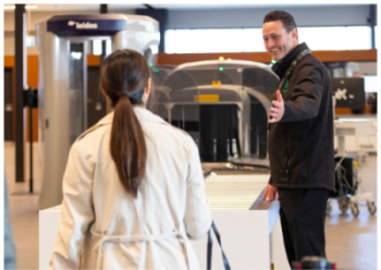A lady in a white coat at the Hobart Airport Security Checkpoint with a security officer directing towards the conveyor belt and scanner. Scanning equipment are visible in the background.