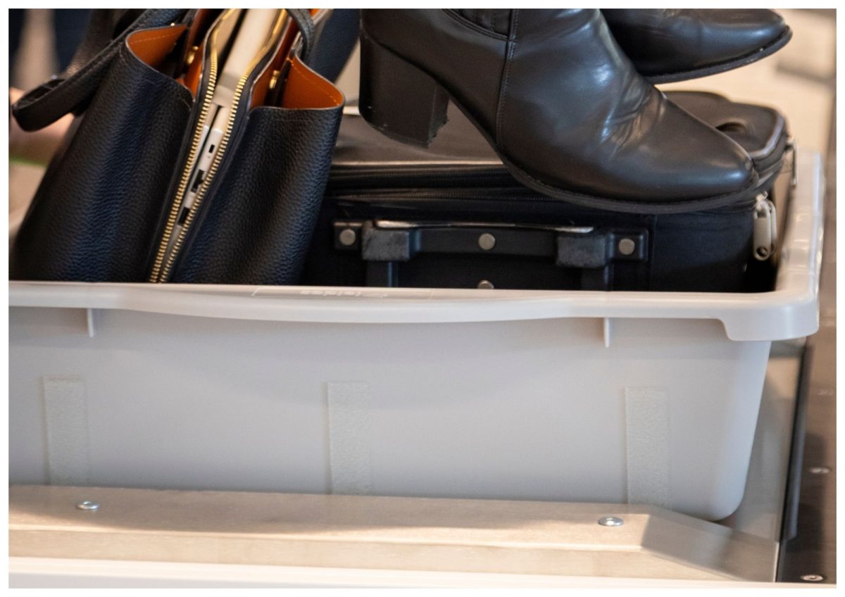 Person placing black ankle boots, a handbag, and a small suitcase into a gray plastic tray on the conveyor belt at the Hobart airport security checkpoint.