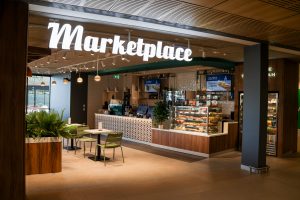 Cafe image with illuminated signage 'Marketplace, tables chairs and a service counter with food and drinks displayed.