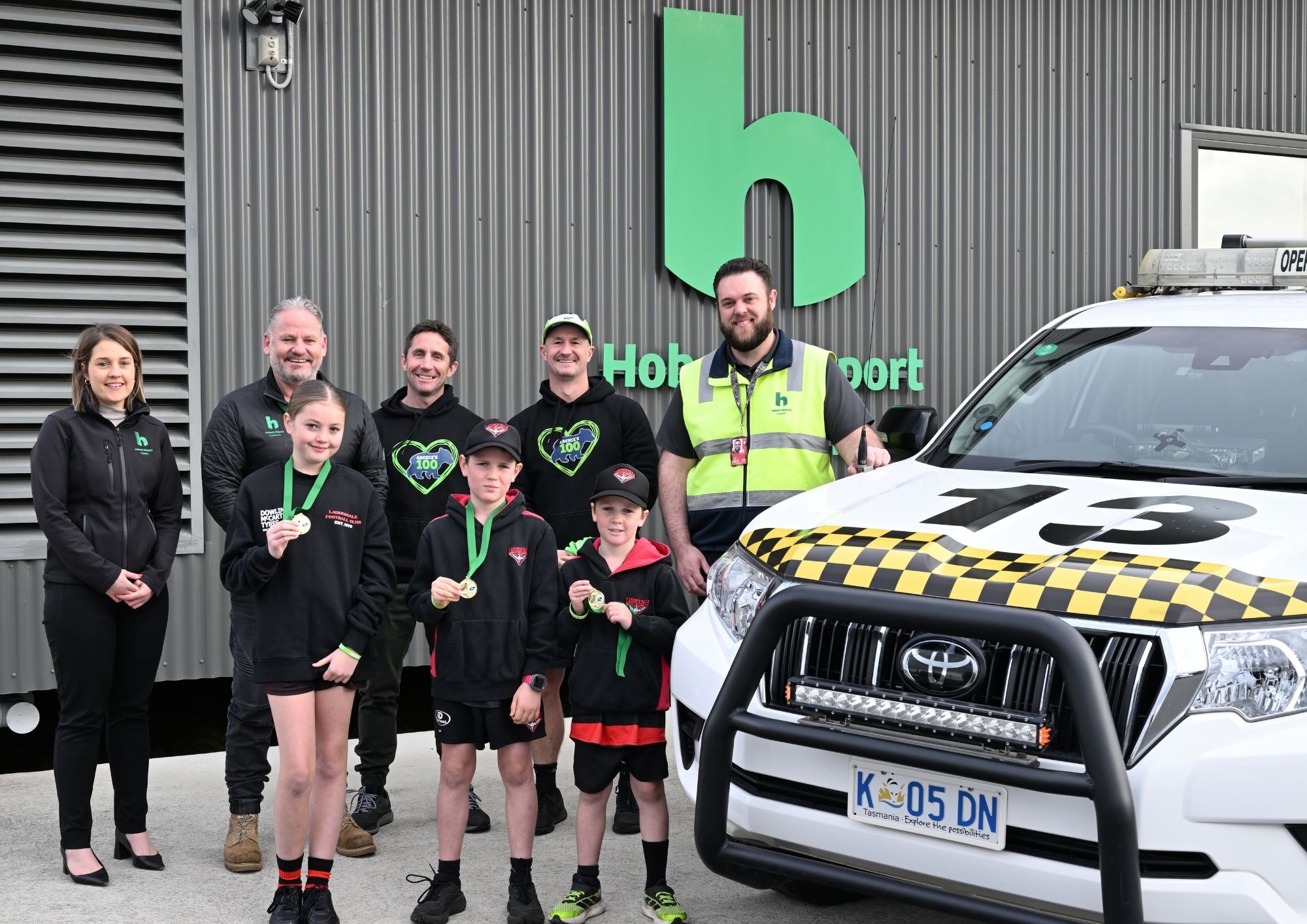 a group of people representing the airport and Archies100 stand together next to an Operations vehicle.
